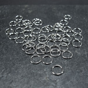 May include: A scattering of small, shiny, silver metal rings on a dark grey surface. The circular rings appear to be made of a smooth, reflective material, possibly stainless steel. They are likely used for jewellery making or crafting.