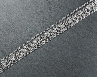 May include: A close-up of multiple silver chains laid out on a dark gray surface. The chains are all the same length and width, and they are arranged in a parallel pattern.