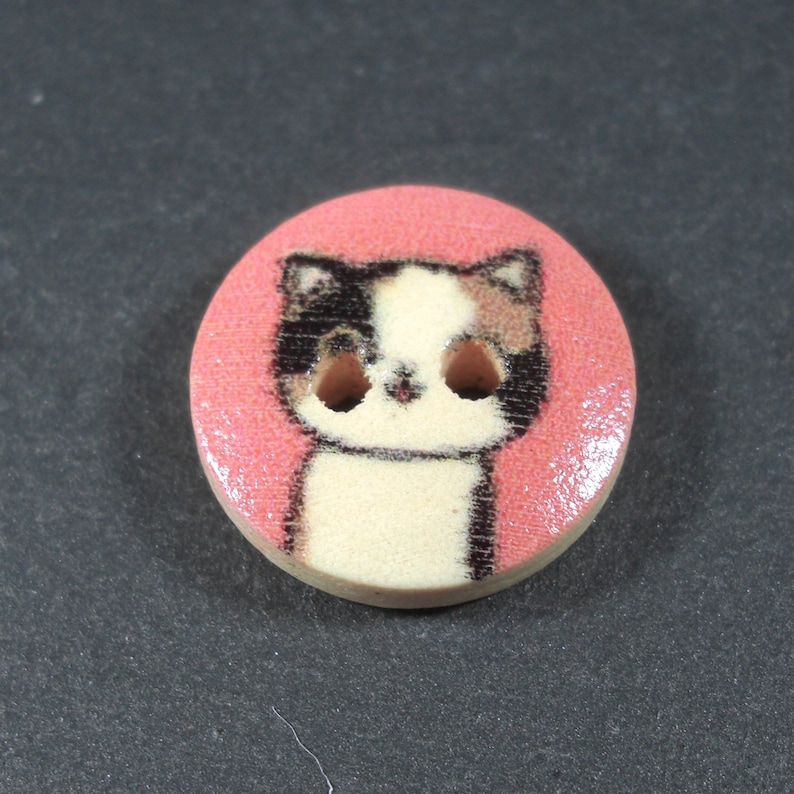 Puede incluir: Un bot&oacute;n rosa con una imagen de dibujos animados de un gato calico con marcas negras y marrones. El gato tiene pelaje blanco y ojos grandes y negros.