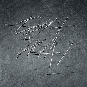 Puede incluir: Primer plano de un montón de alfileres de coser plateados con cabezas redondas esparcidos sobre una superficie gris oscuro.