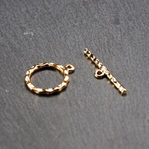 May include: Two gold-toned metal jewellery clasps. One is a round ring with a textured edge. The other is a straight bar with a loop at each end.