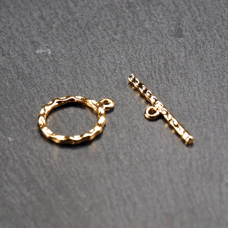 May include: Two gold-toned metal jewellery clasps. One is a round ring with a textured edge. The other is a straight bar with a loop at each end.