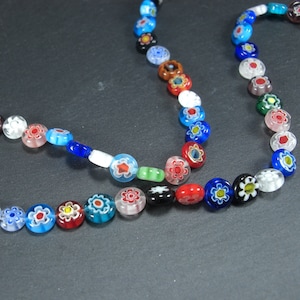 Könnte beinhalten: Eine bunte Halskette aus Glasperlen mit Blumenmustern. Die Perlen sind rund und flach und auf einem durchsichtigen Faden aufgefädelt. Die Halskette ist eine Mischung aus blauen, roten, weißen, grünen und schwarzen Perlen.