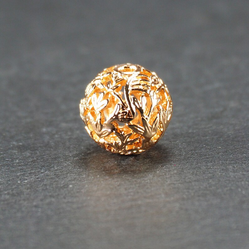 May include: A gold-colored metal bead with a delicate, intricate design. The bead is round and has a hollow center, creating a delicate, airy look.
