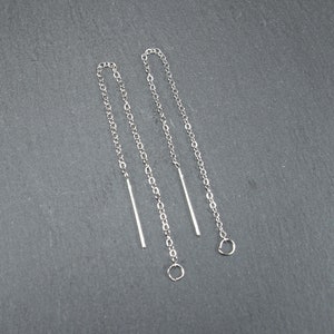 Puede incluir: Un par de pendientes de cadena plateada con un pequeño bucle en el extremo de cada cadena. Las cadenas miden aproximadamente 5 cm de largo y tienen un diseño simple y delicado.