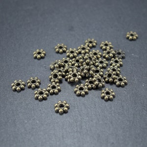 Può includere: Un mucchio di piccole perline di metallo dorate con un design a forma di fiore. Le perline sono sparse su una superficie grigio scuro.