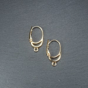 Könnte beinhalten: Zwei goldfarbene Ohrhaken mit einer Schlaufe am unteren Ende. Die Haken sind oval geformt und haben ein doppeltes Drahtdesign.