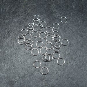 May include: A pile of silver jump rings on a grey background. The rings are small and round, and they are perfect for jewellery making.
