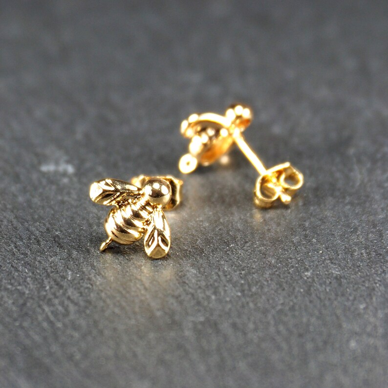 May include: A pair of gold bee stud earrings. The bee is detailed with wings and a segmented body.