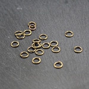 May include: A pile of small, gold-coloured jump rings on a dark grey surface. The rings are about 1.2 cm in diameter.