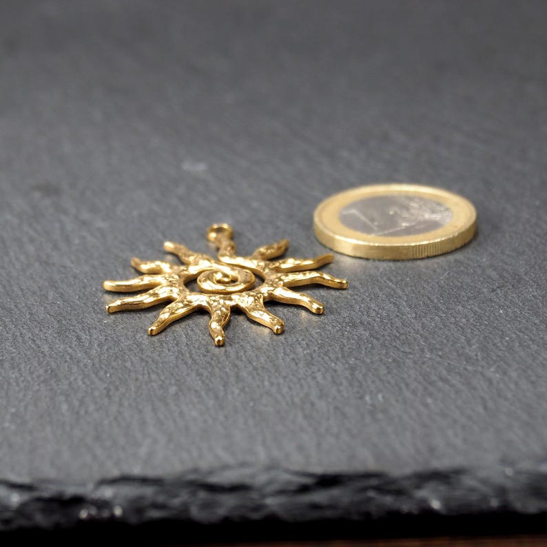 May include: A gold-toned metal sun pendant with a spiral design in the center. The pendant is textured and has a slightly rough surface.