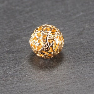 May include: A gold-colored metal bead with a delicate, intricate floral design. The bead is hollow and has a round shape.