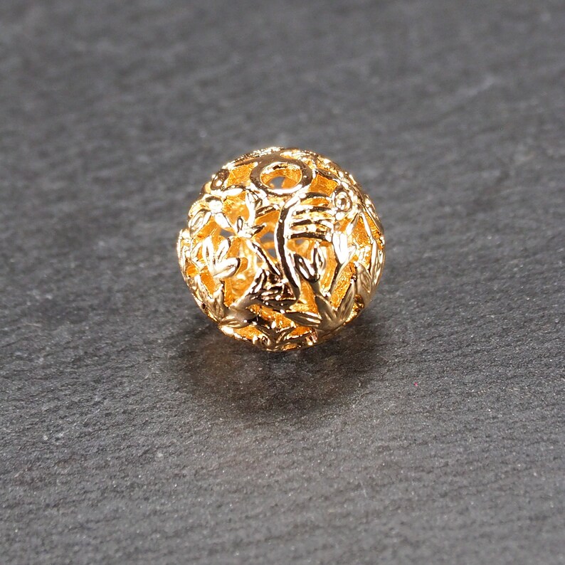 May include: A gold-colored metal bead with a delicate, intricate floral design. The bead is hollow and has a round shape.