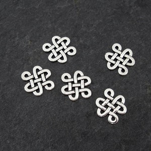 Puede incluir: Seis abalorios de nudo celta de metal plateado, cada uno con forma de estrella de cuatro puntas, dispuestos sobre un fondo gris oscuro.