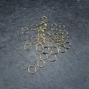 May include: A scattering of small, gold-coloured jump rings on a dark grey surface. The rings are circular and appear to be metal. The lighting highlights their shiny surface, suggesting they are new.