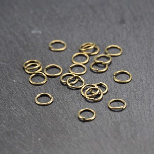 May include: A collection of gold jump rings, scattered on a dark grey surface. These rings are used for jewellery making and crafting.