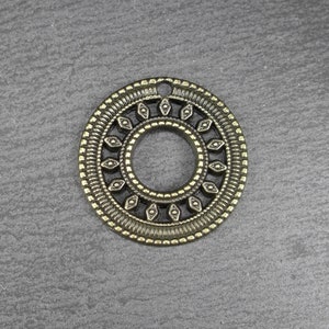 Pode incluir: Um pingente circular de cor bronze com um design intrincado. O pingente apresenta um orifício central rodeado por um padrão de recortes em forma de diamante e bordas externas detalhadas. A estética geral é vintage e ornamentada.