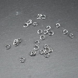 May include: A close-up of a group of silver jump rings, scattered on a dark grey surface. The rings are small and circular, with a smooth, polished finish.