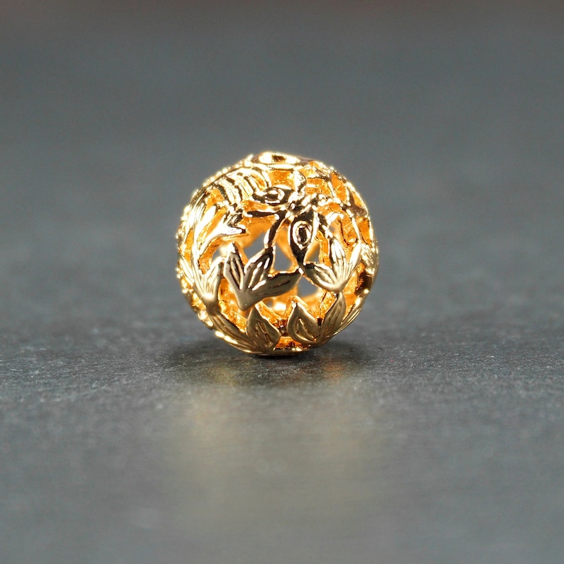 May include: A gold-colored metal bead with a delicate floral design. The bead is hollow and has a round shape.