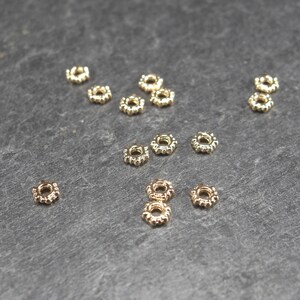 Könnte beinhalten: Eine Gruppe von 14 goldfarbenen Metall-Spacer-Perlen mit einem strukturierten, kreisförmigen Design. Die Perlen sind auf einer grauen Oberfläche verstreut angeordnet.