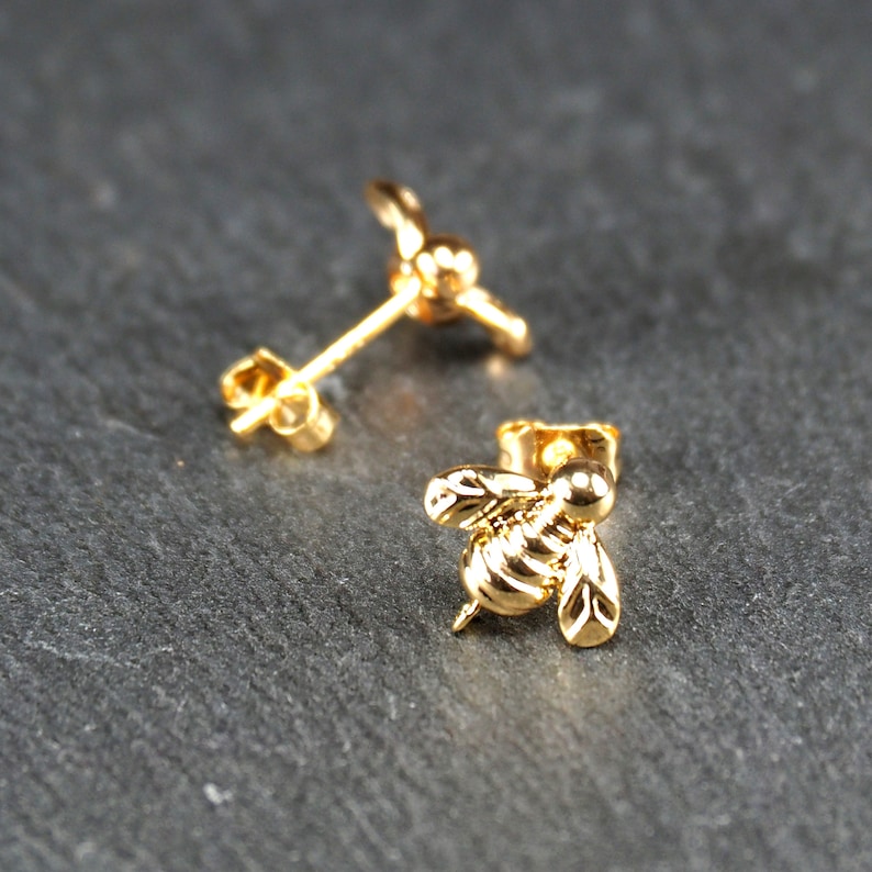 May include: A pair of gold bee stud earrings. The bee is detailed with a textured body and wings.