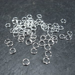 Può includere: Una collezione di piccoli anelli di salto argentati e lucenti sparsi su una superficie grigio scuro. Gli anelli sono circolari con una piccola apertura, probabilmente per la creazione di gioielli o artigianato. Gli anelli sono in metallo riflettente.
