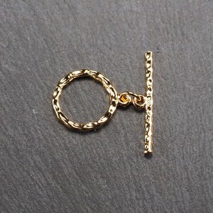 May include: Gold-toned metal toggle clasp with a textured finish. The clasp has a round ring and a straight bar.