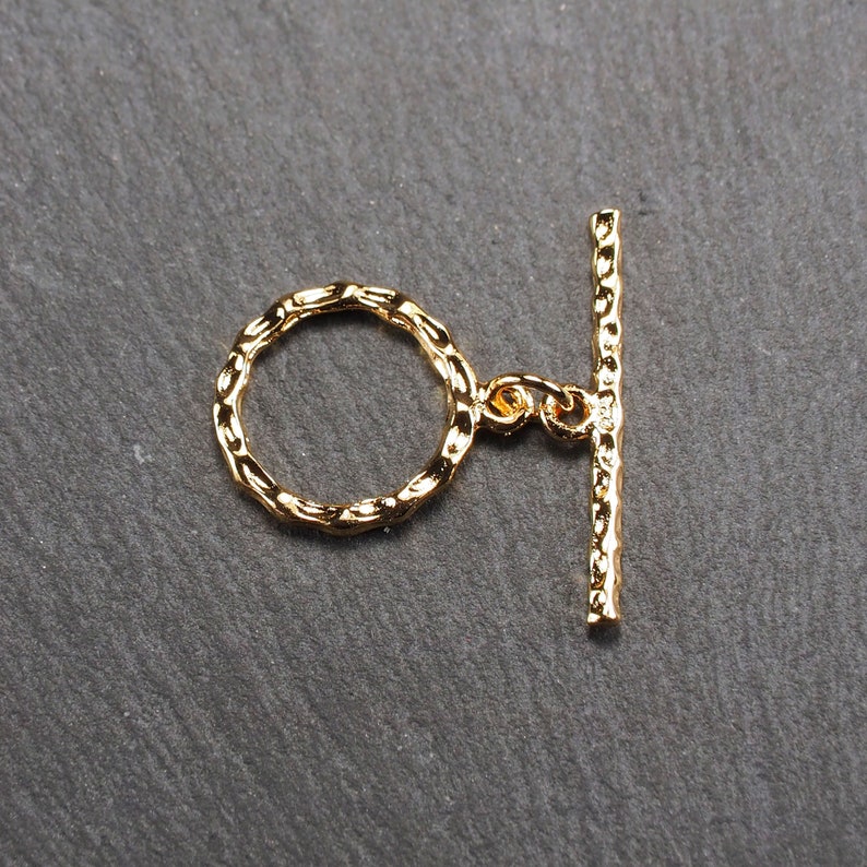 May include: Gold-toned metal toggle clasp with a textured finish. The clasp has a round ring and a straight bar.