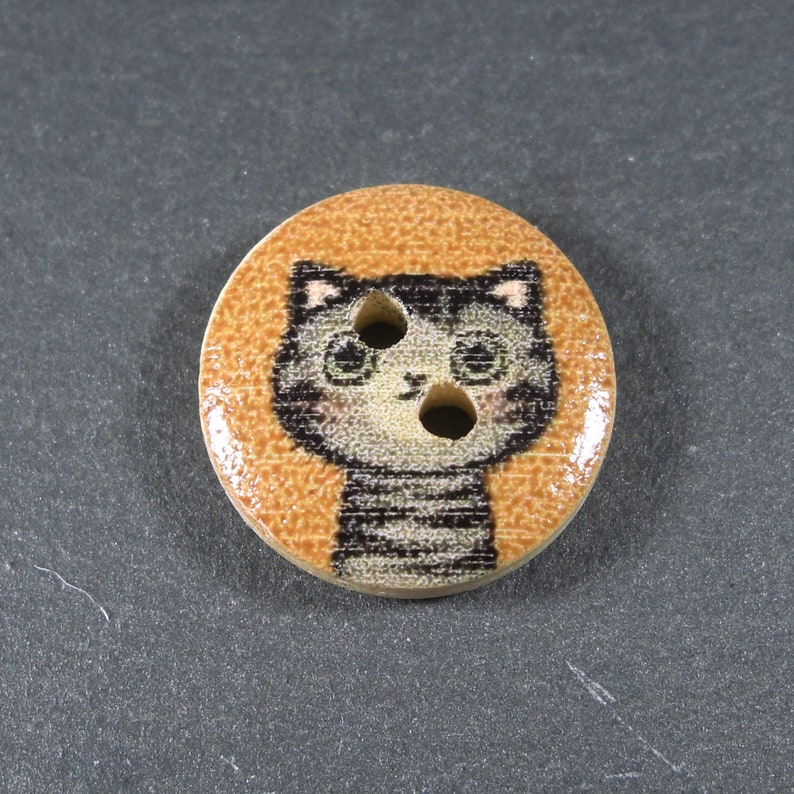Puede incluir: Un bot&oacute;n de madera con fondo marr&oacute;n y un gato de dibujos animados en blanco y negro con un patr&oacute;n a rayas. El gato tiene dos ojos negros y una nariz rosa.
