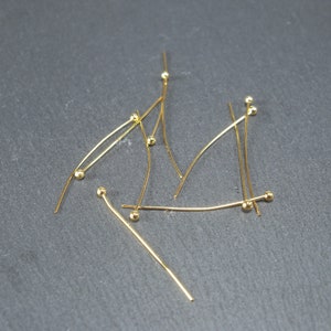 May include: A group of gold-coloured head pins with round heads. The pins are arranged in a random pattern on a grey surface.