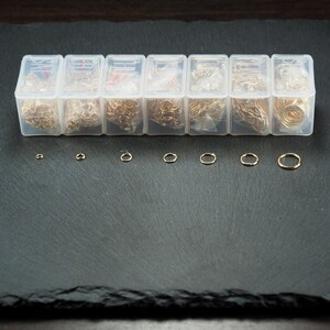 以下が含まれることがあります： さまざまなサイズの金色のジャンプリングが詰まった透明なプラスチック容器が8個セットになっています。容器は黒いスレートの表面に一列に並んでいます。容器の下には、さまざまなサイズの金色のジャンプリングが8個並んでいます。