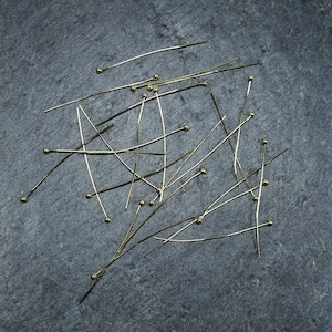 May include: A close-up of a group of gold-coloured straight pins with round heads scattered on a dark grey surface.