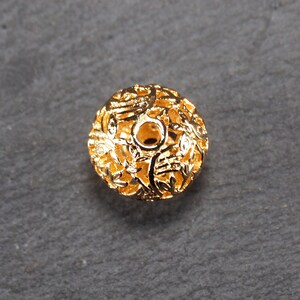 May include: A gold-colored metal bead with a delicate, intricate design. The bead is hollow and has a round shape.