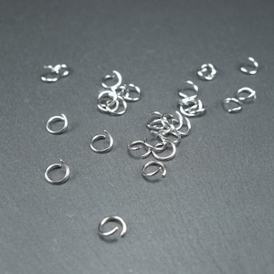 Può includere: Primo piano di un mucchio di anelli di salto argentati, un tipo di componente di gioielleria utilizzato per collegare catene, cordini e altri componenti di gioielleria. Gli anelli sono sparsi su una superficie grigio scuro.