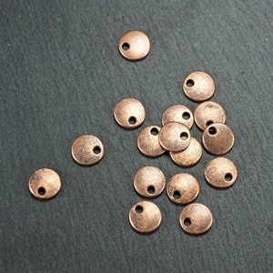 Könnte beinhalten: Eine Gruppe von 14 kupferfarbenen Metallkreisen mit einem kleinen Loch in der Mitte. Die Kreise sind in einer Gruppe auf einer dunkelgrauen Oberfläche angeordnet.