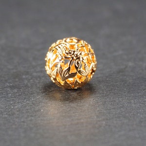 May include: A gold-colored metal bead with a delicate floral design. The bead is round and has a hollow center.