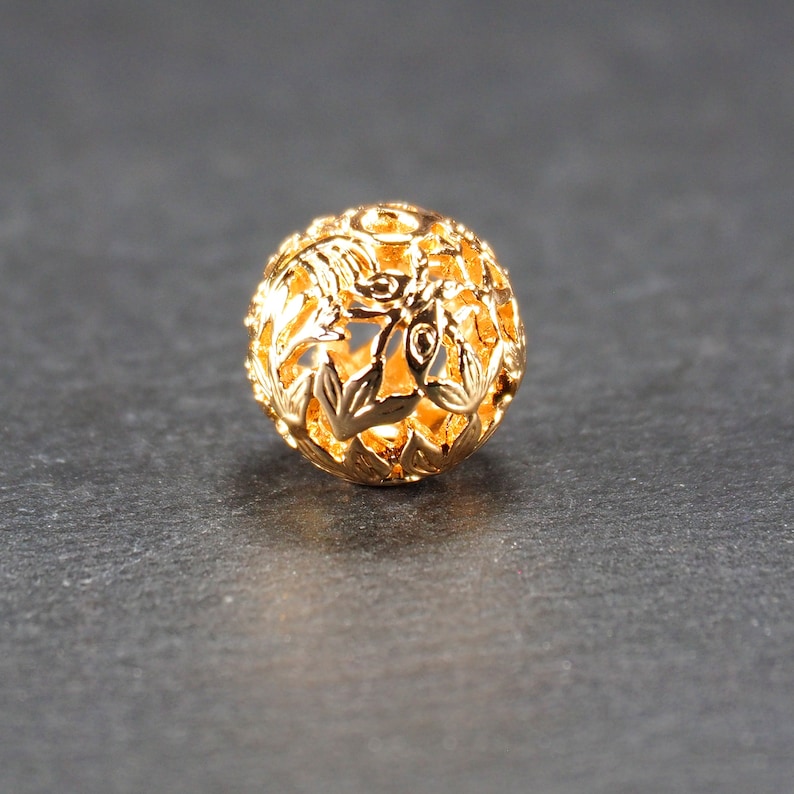 May include: A gold-colored metal bead with a delicate floral design. The bead is round and has a hollow center.