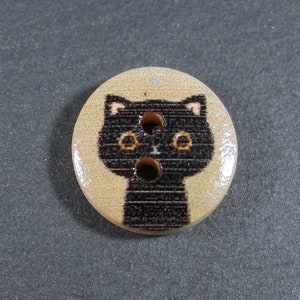 Puede incluir: Un bot&oacute;n de madera con una cara de gato negro impresa. El gato tiene ojos amarillos y una nariz rosa.