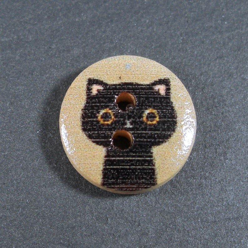 Puede incluir: Un bot&oacute;n de madera con una cara de gato negro impresa. El gato tiene ojos amarillos y una nariz rosa.