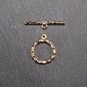 May include: Gold-toned metal toggle clasp with a textured finish. The clasp features a round ring and a straight bar with a loop at each end.