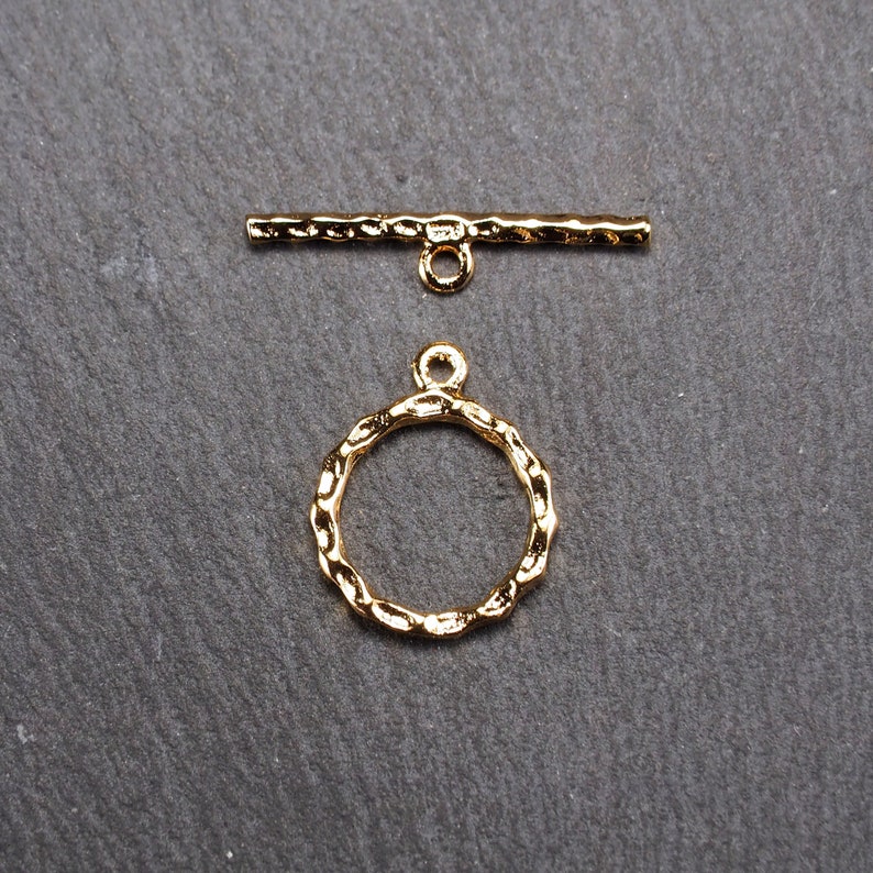 May include: Gold-toned metal toggle clasp with a textured finish. The clasp features a round ring and a straight bar with a loop at each end.
