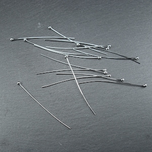 May include: A pile of silver head pins with round heads. The pins are scattered on a dark grey surface.