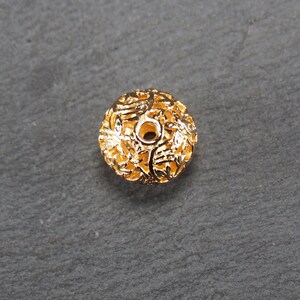 May include: Gold-toned metal bead with a delicate, intricate floral design. The bead has a round shape and a hole in the center for stringing.