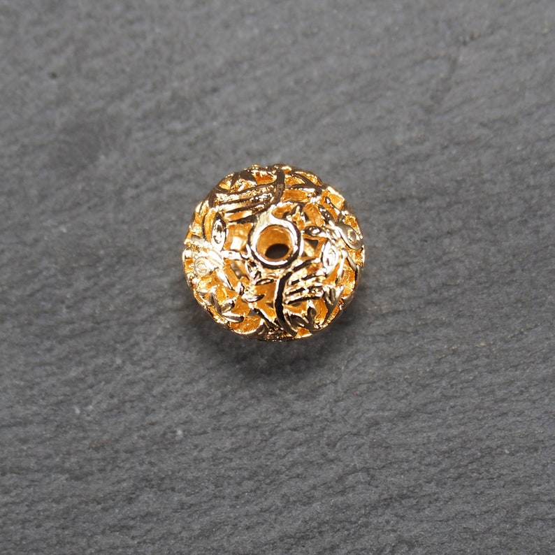 May include: Gold-toned metal bead with a delicate, intricate floral design. The bead has a round shape and a hole in the center for stringing.