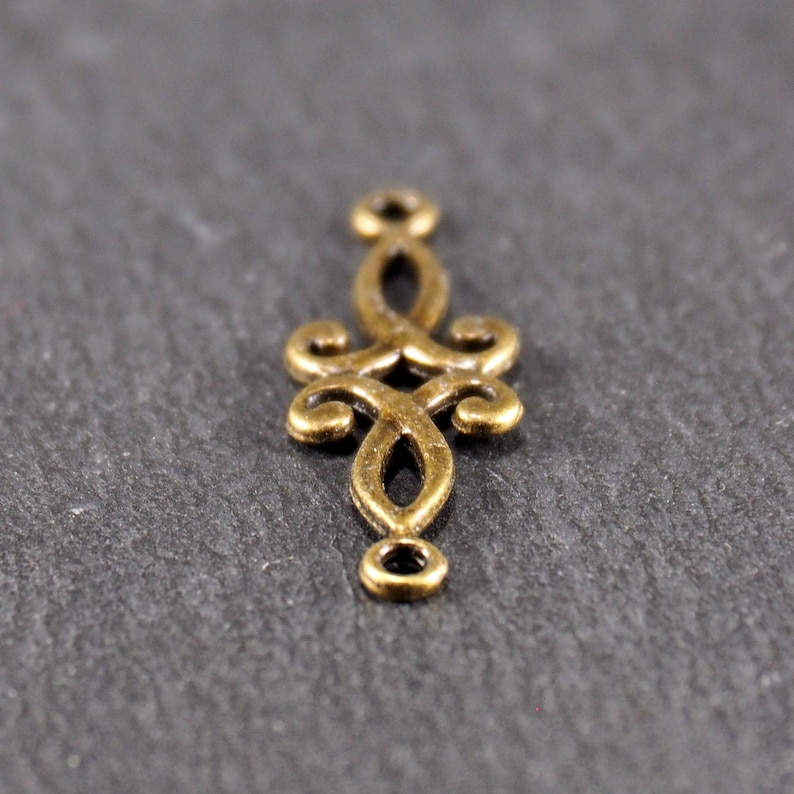 Puede incluir: Un conector de metal dorado con un dise&ntilde;o decorativo y ornamentado. El conector tiene dos bucles en la parte superior e inferior, lo que lo hace ideal para la elaboraci&oacute;n de joyas u otros proyectos de artesan&iacute;a.