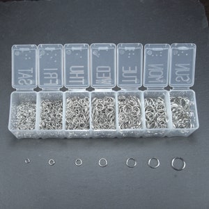 Op de afbeelding: Een transparante plastic opbergdoos met zeven vakken, elk gevuld met zilveren springringen in verschillende maten. De doos is gelabeld met de dagen van de week: Zo, Ma, Di, Wo, Do, Vr, Za.  Onder de doos bevinden zich zeven zilveren springringen in verschillende maten.