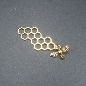 Pode incluir: Encanto de metal dourado com um design de favo de mel e um pequeno pingente de abelha.