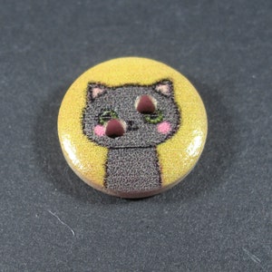 Puede incluir: Un bot&oacute;n redondo amarillo con un gato gris de dibujos animados con mejillas rosadas y ojos verdes. El gato est&aacute; mirando hacia arriba con una expresi&oacute;n ligeramente sorprendida.