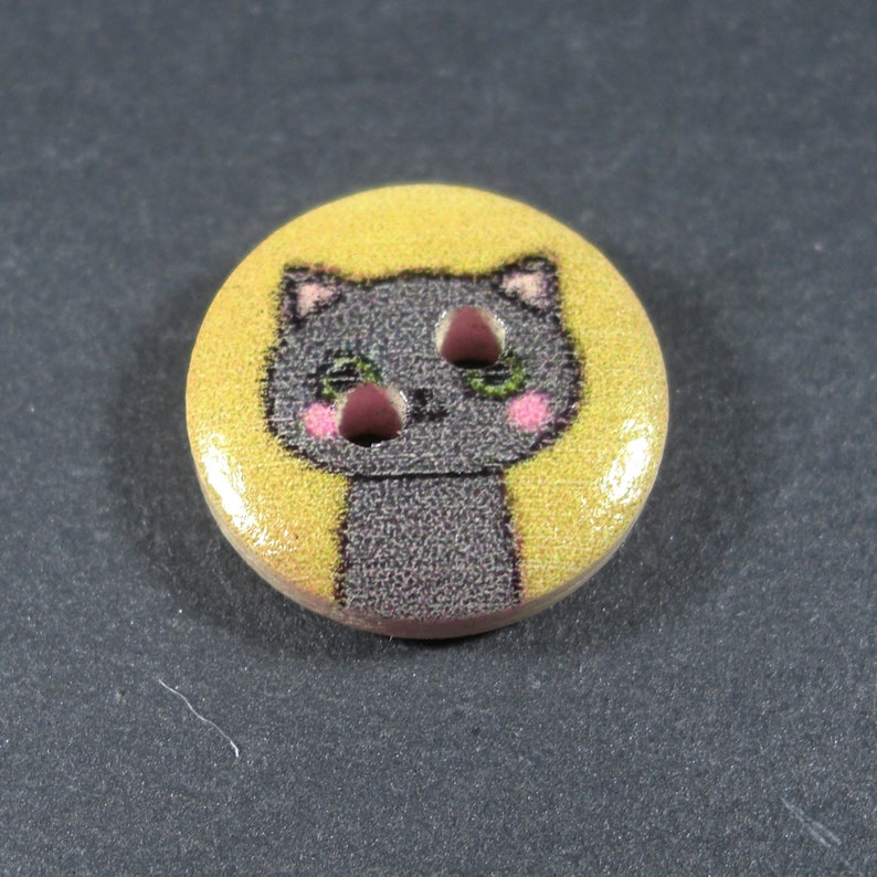 Puede incluir: Un bot&oacute;n redondo amarillo con un gato gris de dibujos animados con mejillas rosadas y ojos verdes. El gato est&aacute; mirando hacia arriba con una expresi&oacute;n ligeramente sorprendida.