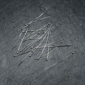 Puede incluir: Un montón de alfileres rectos plateados con cabezas redondas esparcidos sobre una superficie gris oscuro.